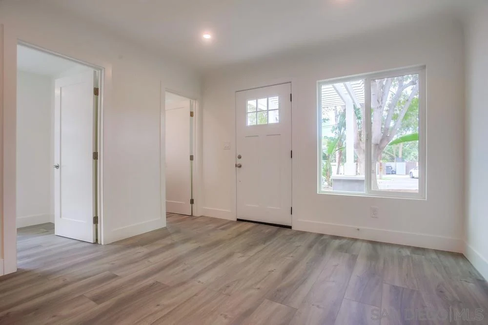 Room with light walls, wood-like floor, front door with window, large window, and two doorways.