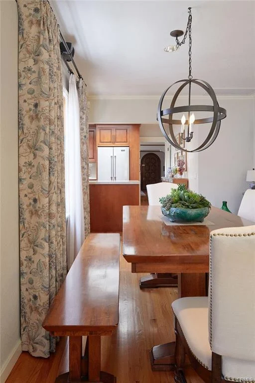 Dining area with wooden table, white chairs, bench, floral curtains, and kitchen in background.