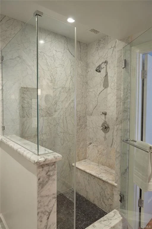 Bathroom shower with marble walls, glass enclosure, bench, and polished fixtures.
