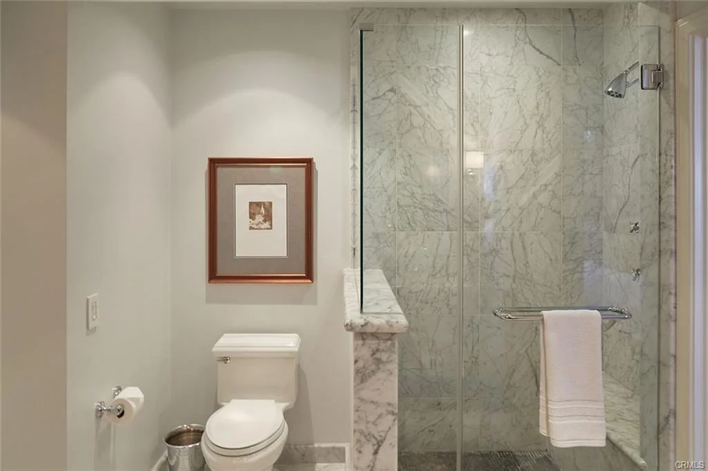 Bathroom with toilet, glass shower, marble walls, and framed picture.