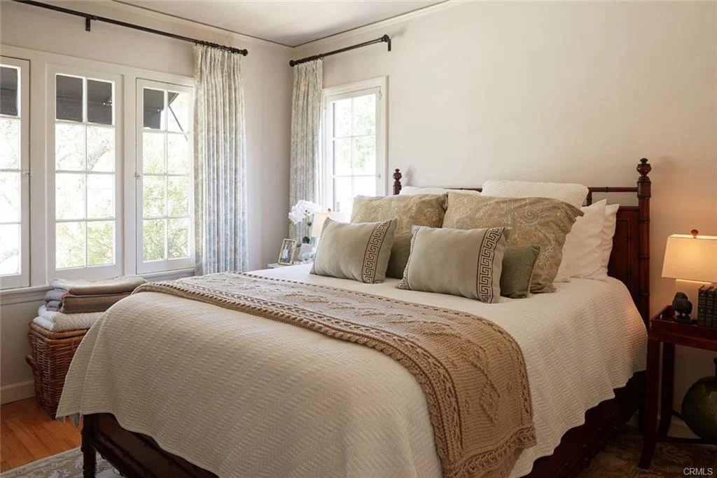 Bedroom with bed, pillows, blanket, windows with curtains, lamp, and basket.