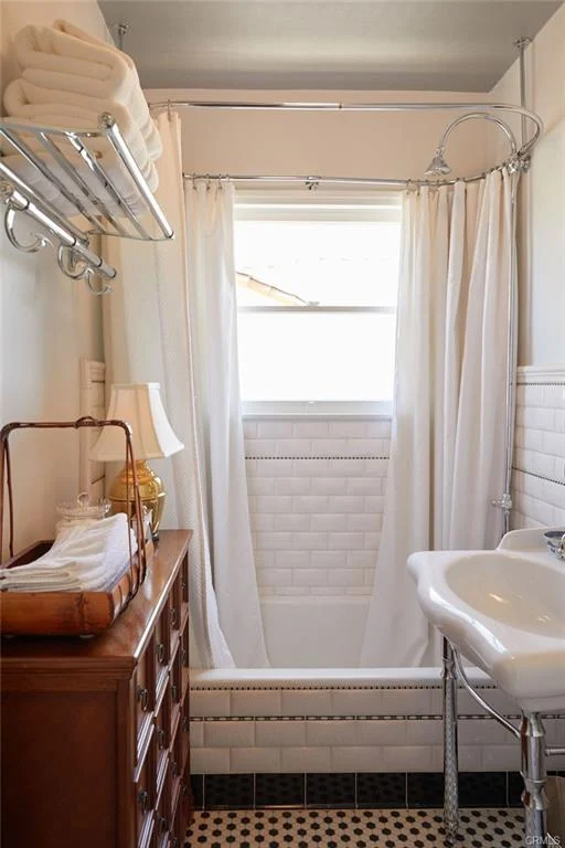 Small bathroom with shower, white curtain, sink on metal stand, towels on shelves, wooden dresser, and patterned floor.