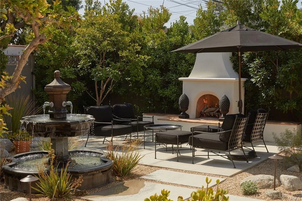 Outdoor area with a fountain, black chairs, umbrella, fireplace, and green plants.
