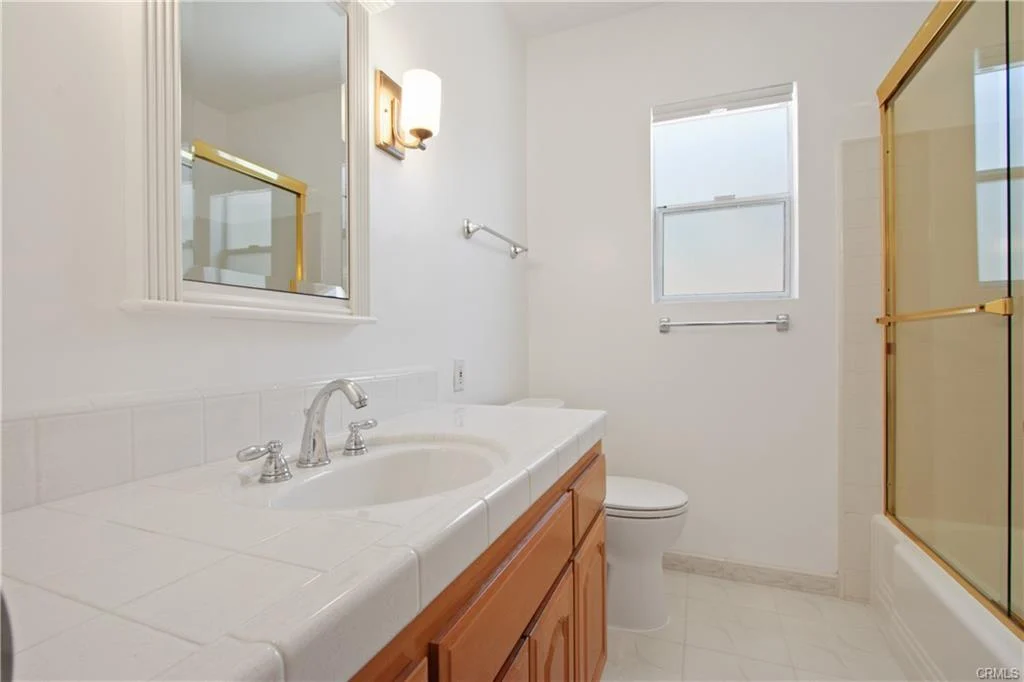 Bathroom with white tile countertop, wooden cabinets, toilet, and glass shower
