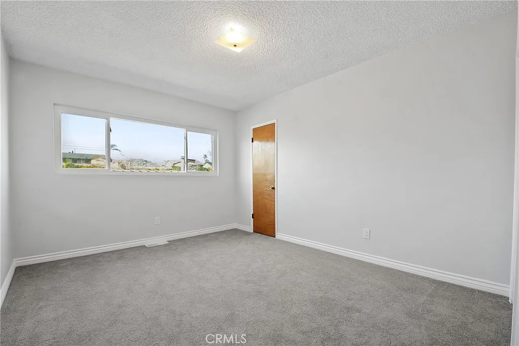 Empty room with white walls, gray carpet, a wood door, and a big window.