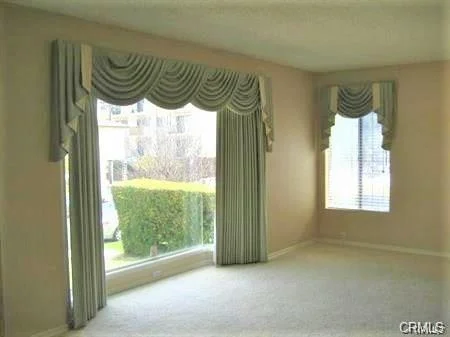 Room with beige walls, carpet, large windows, patterned drapes, and hedge outside